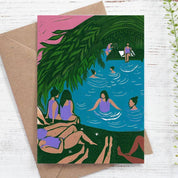 Card with a tropical scene of people by a pool, featuring a brown envelope on a white wooden surface.